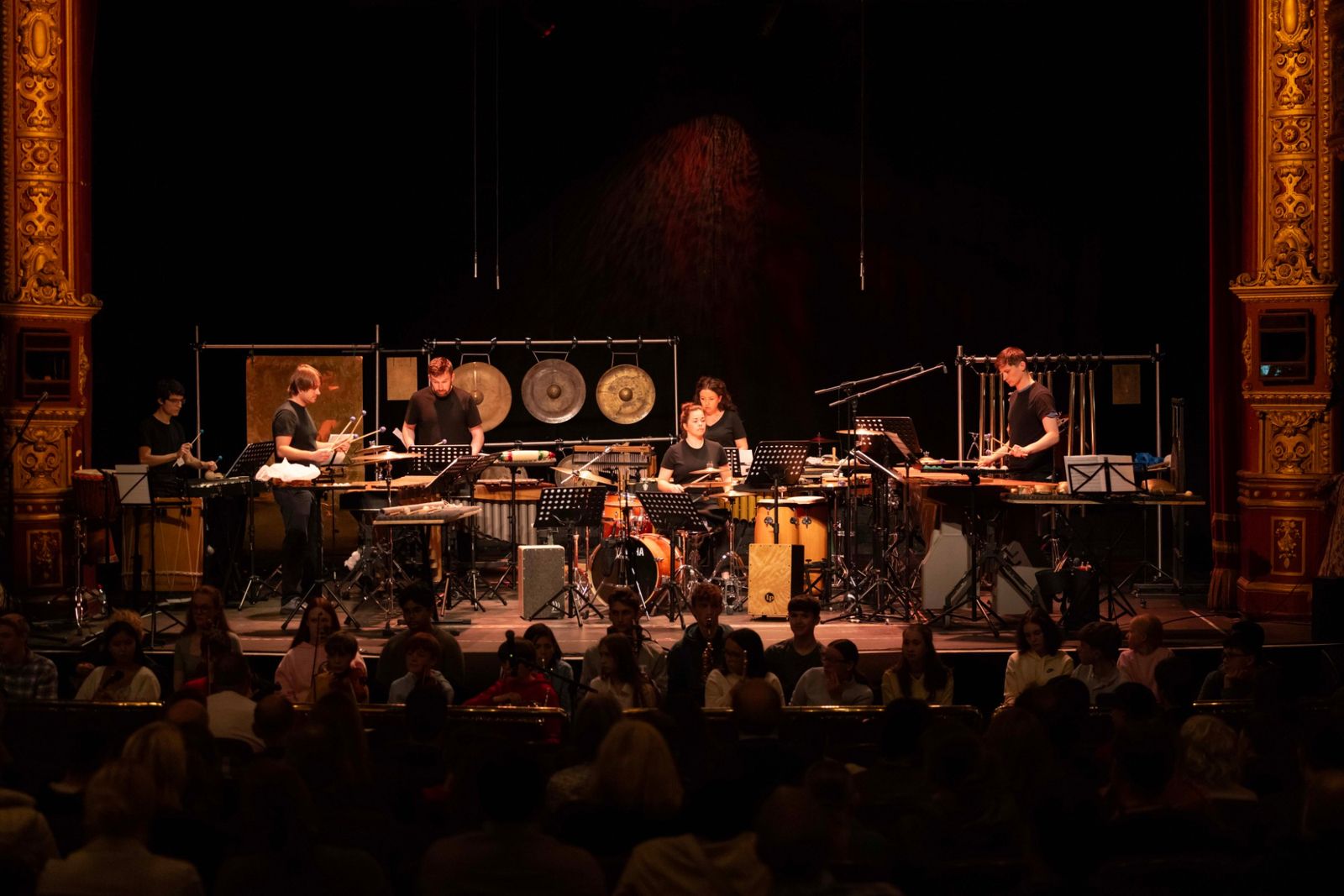 Bangers and Crash on an ornate theatre stage, among gongs, marimbas and drums, with a young audience in front