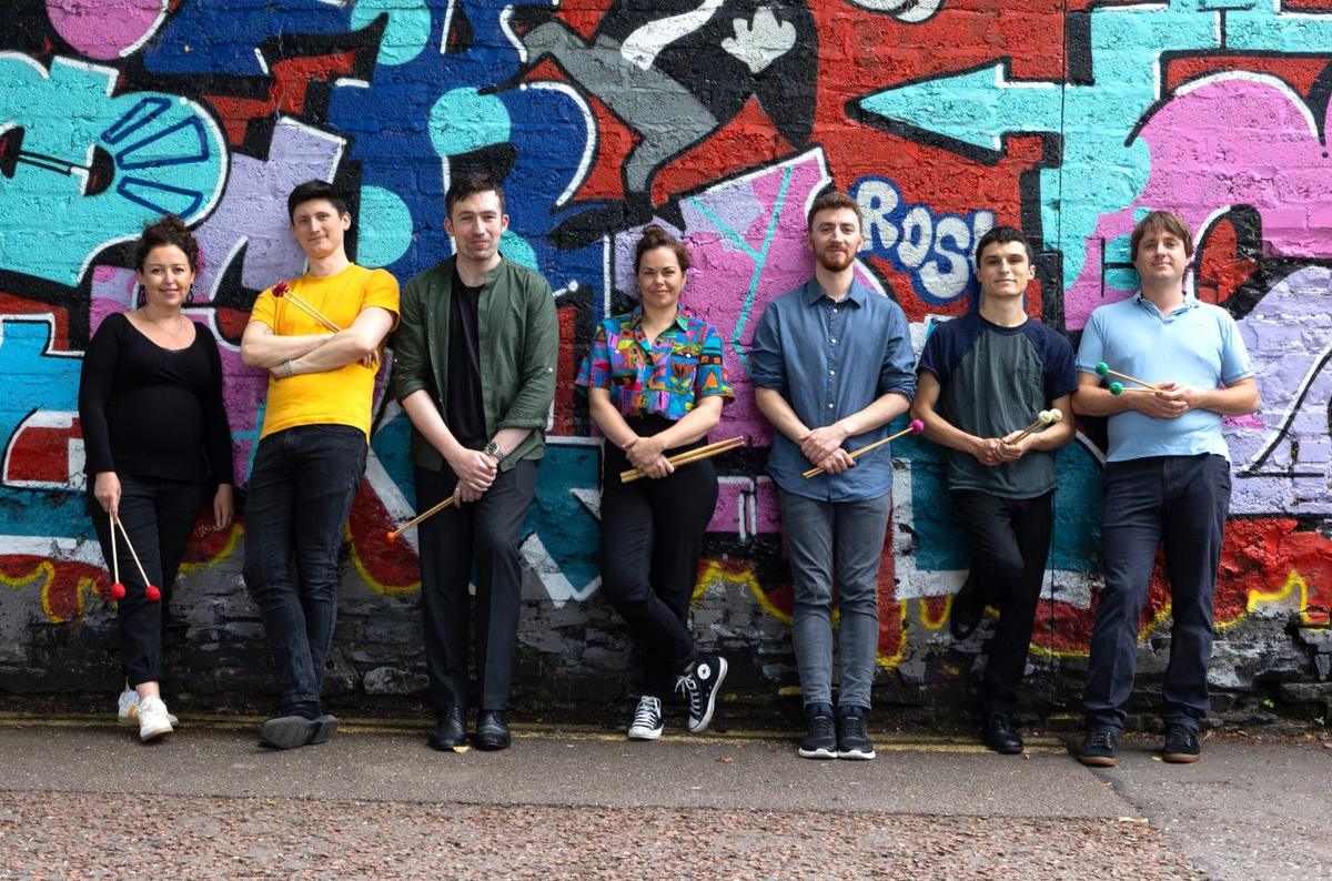 Seven members of Bangers and Crash lined up against a graffiti wall, holding mallets and sticks