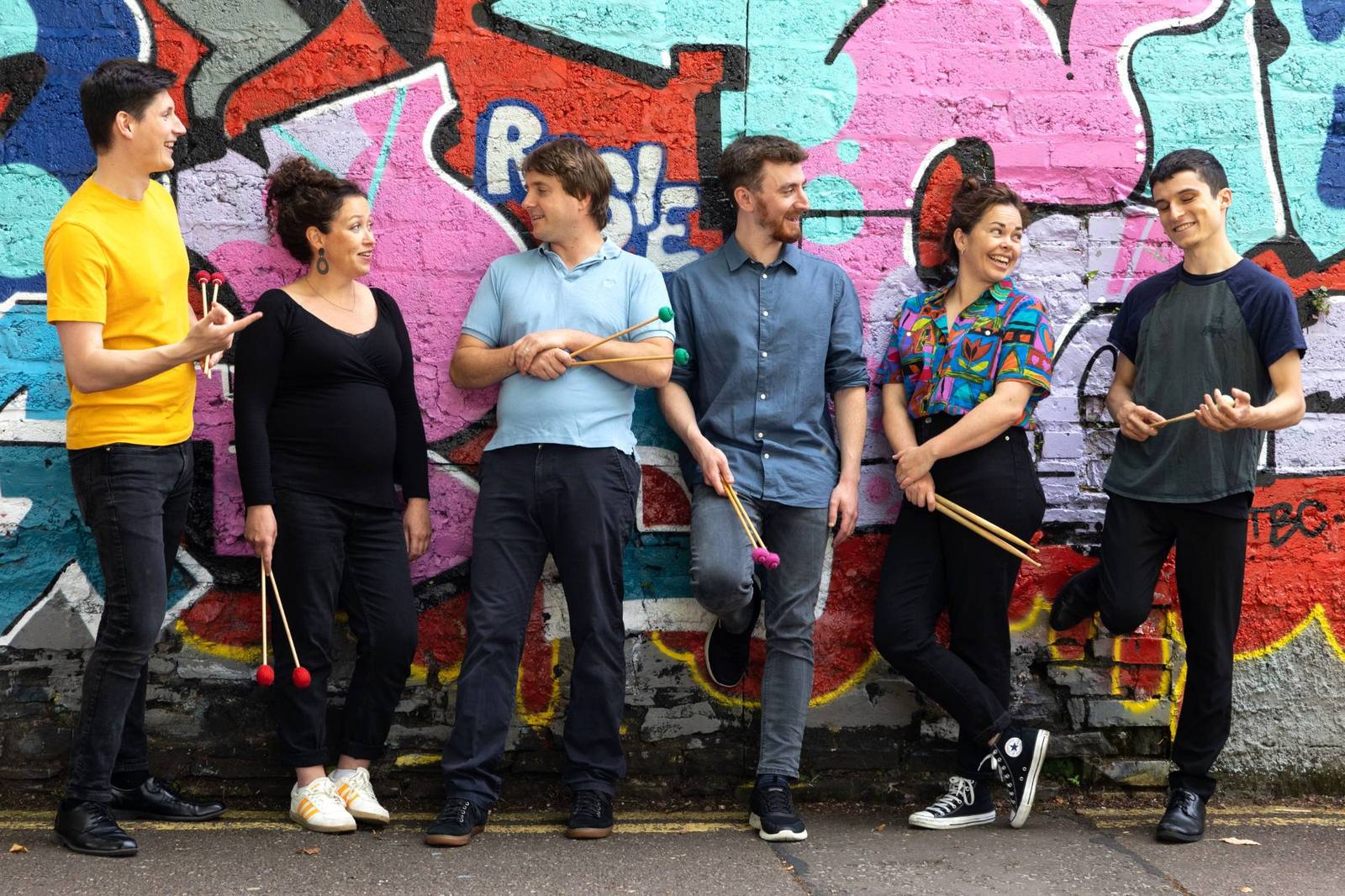 Six members of Bangers and Crash, holding mallets and sticks and laughing, in front of a colourful graffiti wall