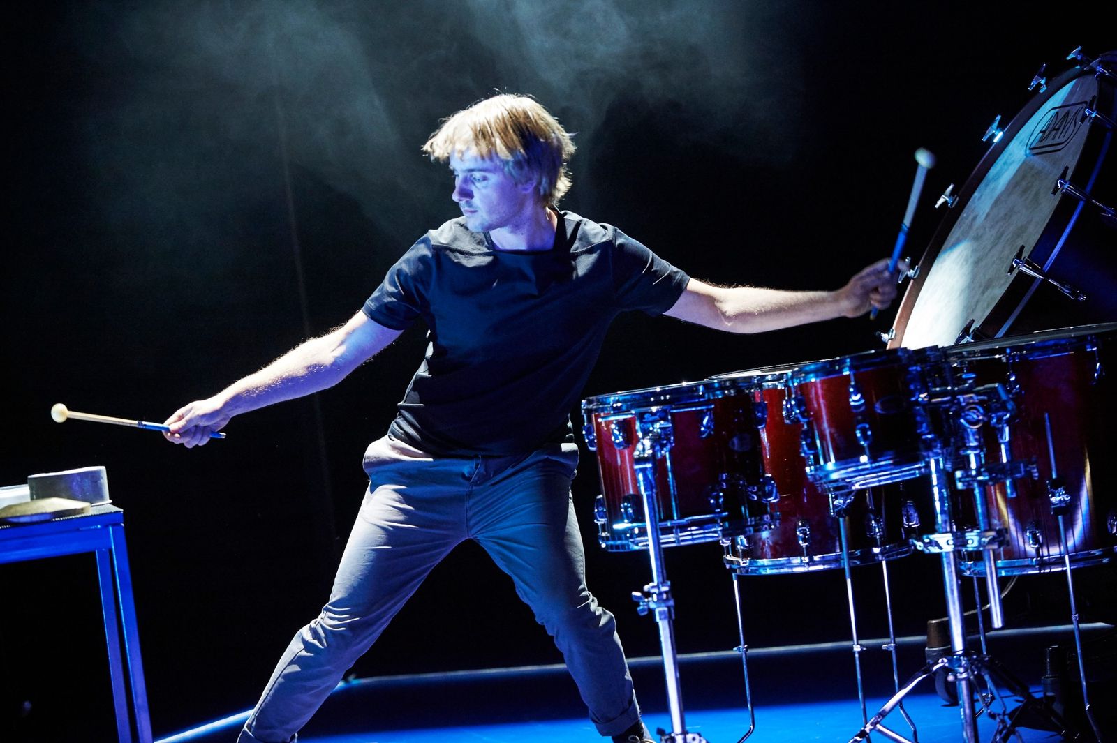 Alex Petcu on a dark stage lit in blue, arms stretched out with a mallet in each hand, between a table of small instruments and a set of red drums and a bass drum